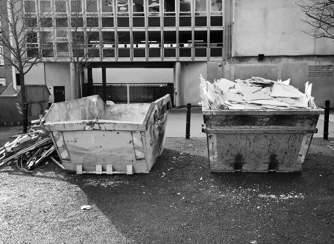 Two large industrial rubbish skips are positioned side by side on a paved surface in front of a modern, grey concrete building with a grid-patterned facade. The left skip contains large, broken pieces of concrete and debris, with rough, uneven textures and a weathered, light grey appearance. The right skip is filled with a considerable amount of crumpled, white waste paper and cardboard, stacked haphazardly, with some edges protruding over the rim. Surrounding the skips, small scattered bits of waste, gravel, and dust are visible on the ground. In the background, metal railings and a partial view of a storage area beneath the building are present. The scene appears to be a typical outdoor waste collection point, relevant to rubbish removal services in Chelmsford, suggesting a typical municipal or commercial waste disposal setting. The lighting is neutral, with no harsh shadows or bright highlights, providing a clear view of the materials and their textures, suitable for describing rubbish clearance activities.