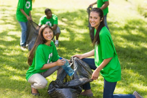 Two young women wearing bright green T-shirts with a white recycling symbol on the front are kneeling on a grassy area, actively collecting waste. They are holding a large black rubbish bag between them, with one woman placing an item inside while the other stabilizes the bag. The women have cheerful expressions and are surrounded by more volunteers in the background, all dressed similarly in green T-shirts, engaged in a community cleanup. The scene is set outdoors on a sunny day, with lush green grass and trees providing a natural backdrop, indicating an organized environmental activity possibly in a local park or community area in Chelmsford, Essex. The overall atmosphere reflects a coordinated effort to promote waste management and recycling, relevant to the services offered by House Clearance Chelmsford in rubbish removal and environmental cleanup activities.