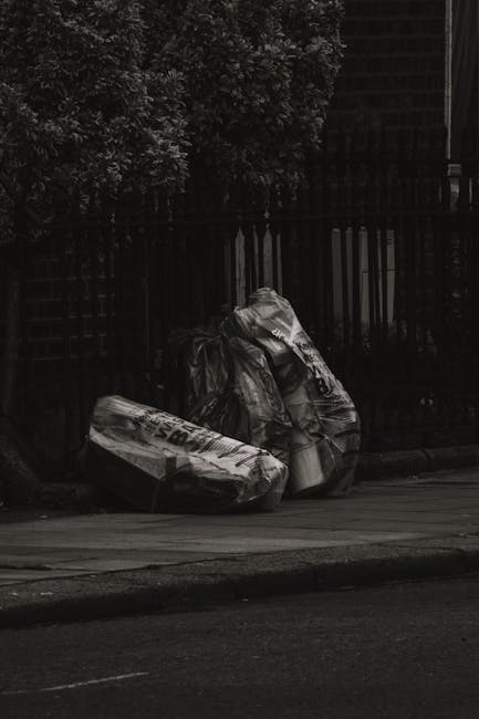 Two large black plastic rubbish bags filled with waste are placed on the pavement beside a black metal fence with vertical bars. The bags appear to be crumpled and loosely tied at the top, with some v